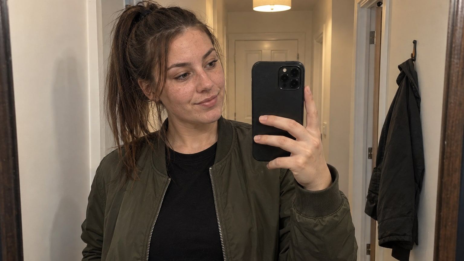 Woman with a high chestnut ponytail takes a mirror selfie in an olive bomber jacket.