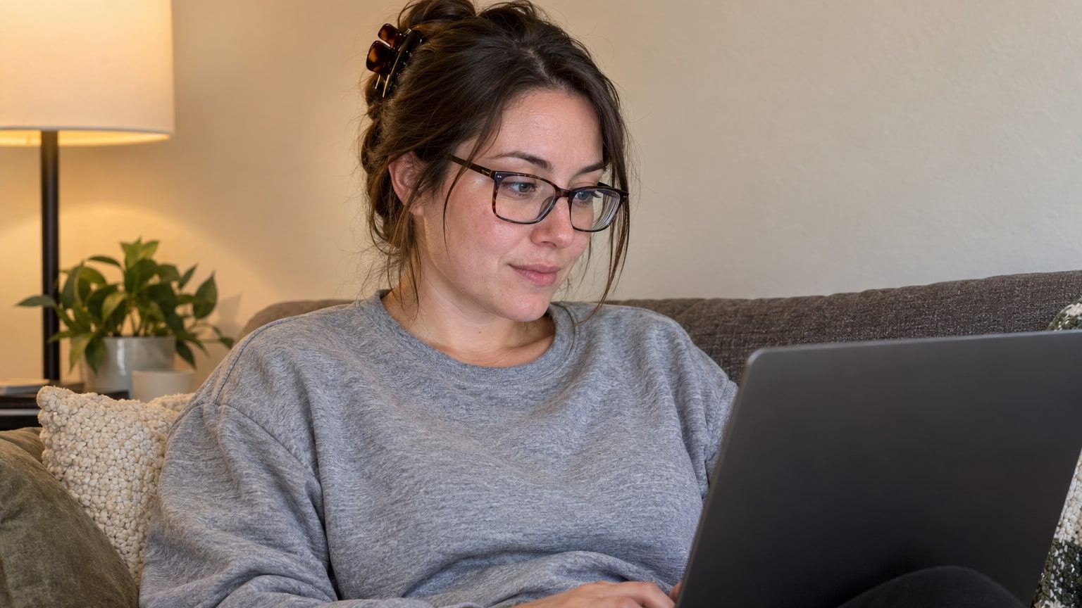 Woman in glasses and a grey sweatshirt reads on a laptop on her couch.