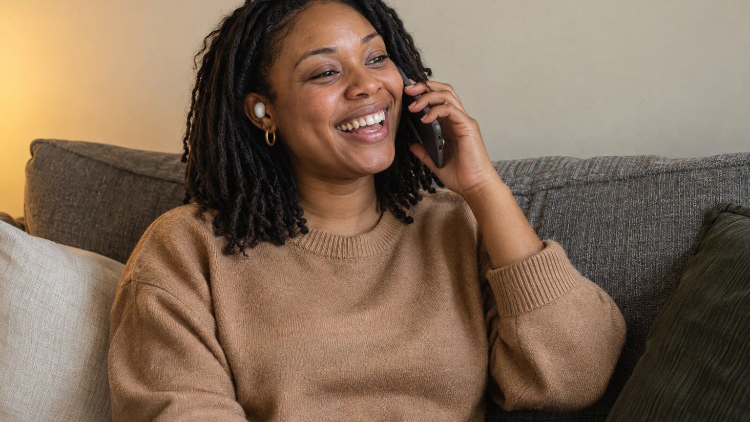 Woman with shoulder-length locs laughs during a phone call on her sofa.