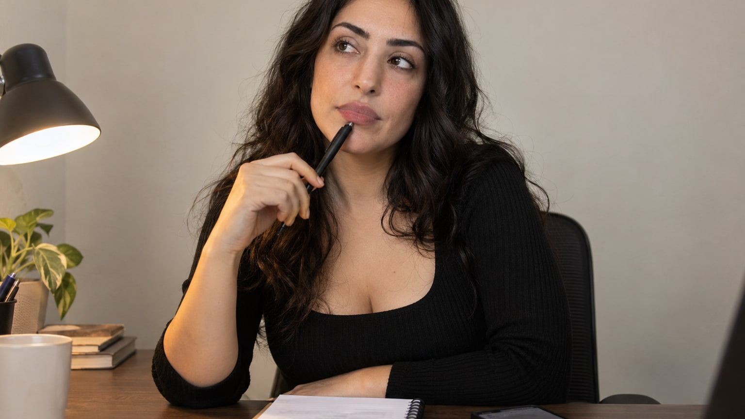 Woman with dark wavy hair taps a pen against her chin while deciding something.