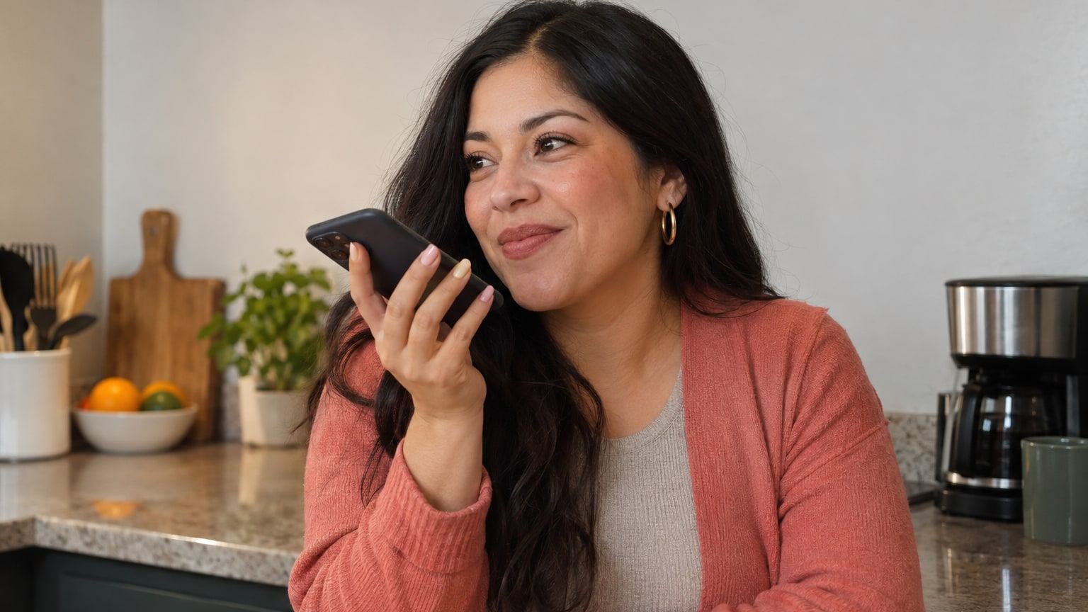 Woman with long dark hair records a voice note with her phone in the kitchen.
